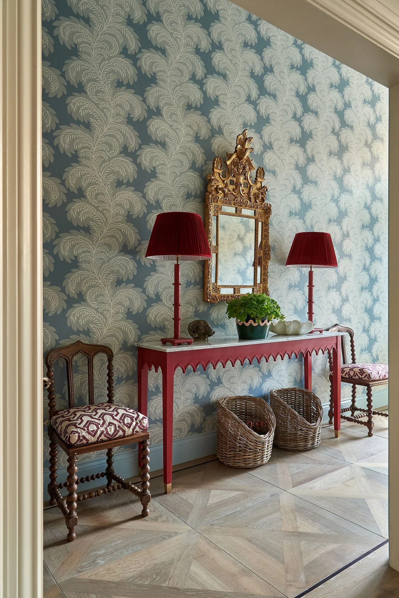Red console table and matching lamps against blue patterned wallpaper beneath an ornate gold-colored mirror.