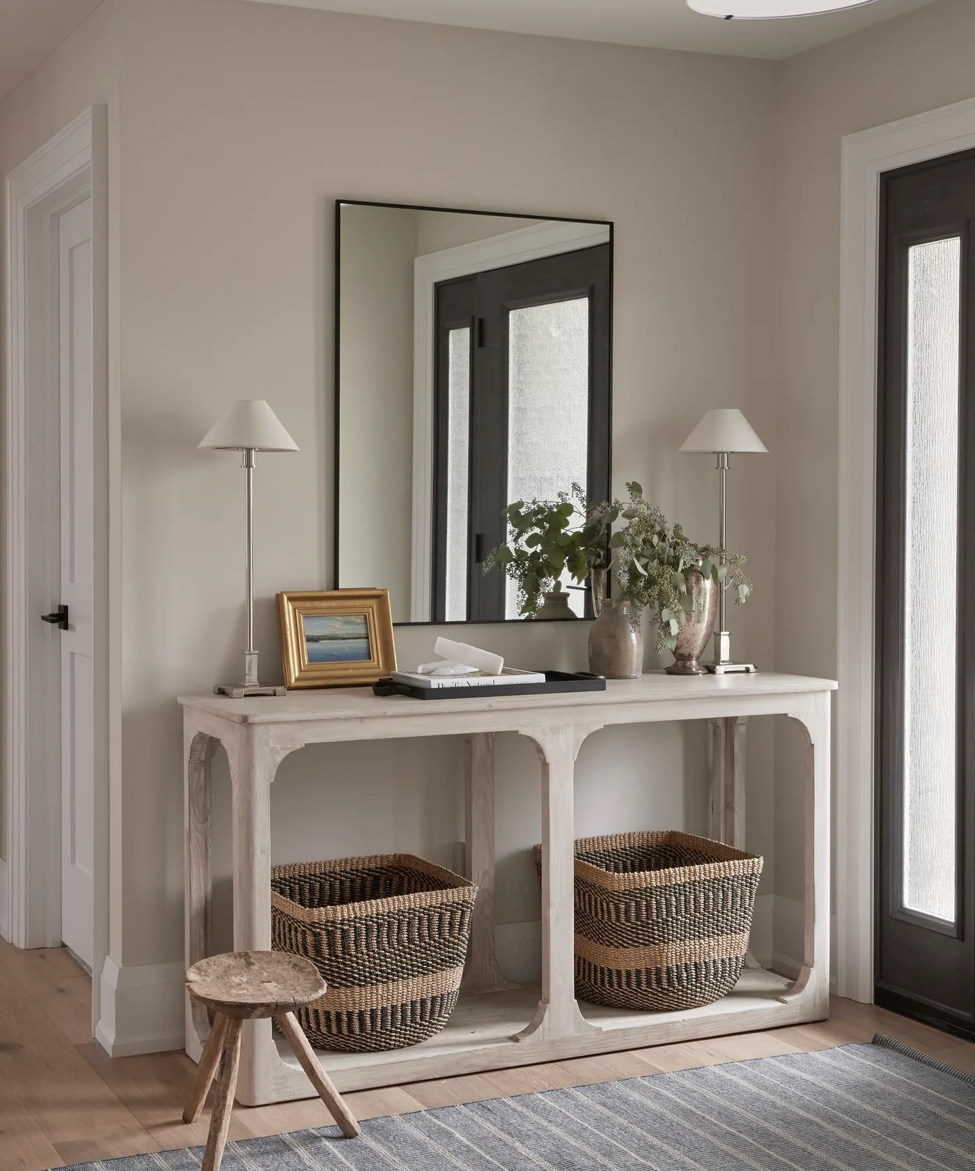 Pale console with large dark-edged mirror, two slim lamps, framed picture, tray, greenery and two striped woven baskets below