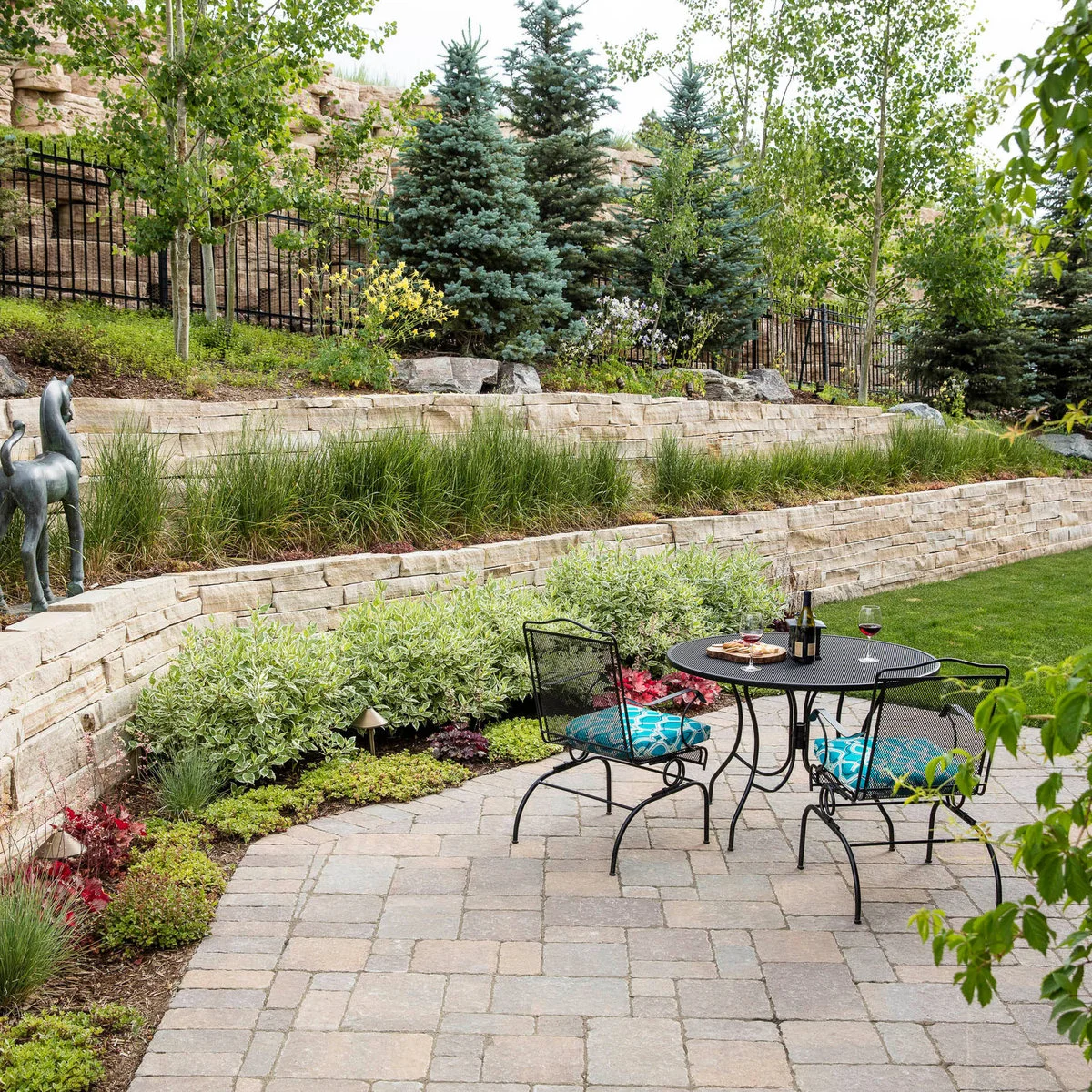 A round two-person patio table sits in front of curved pale stone retaining walls and layered planting.