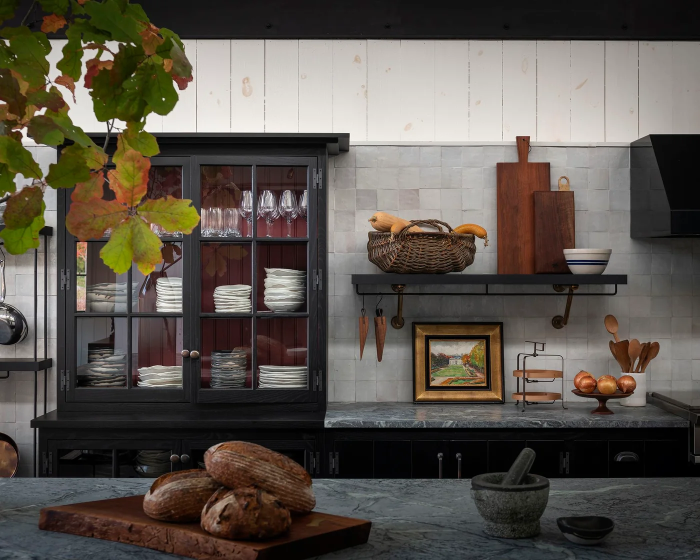 A dark glass-front cabinet displays white dishes beside pale square tiles, a small shelf, framed artwork and a darker veined working surface