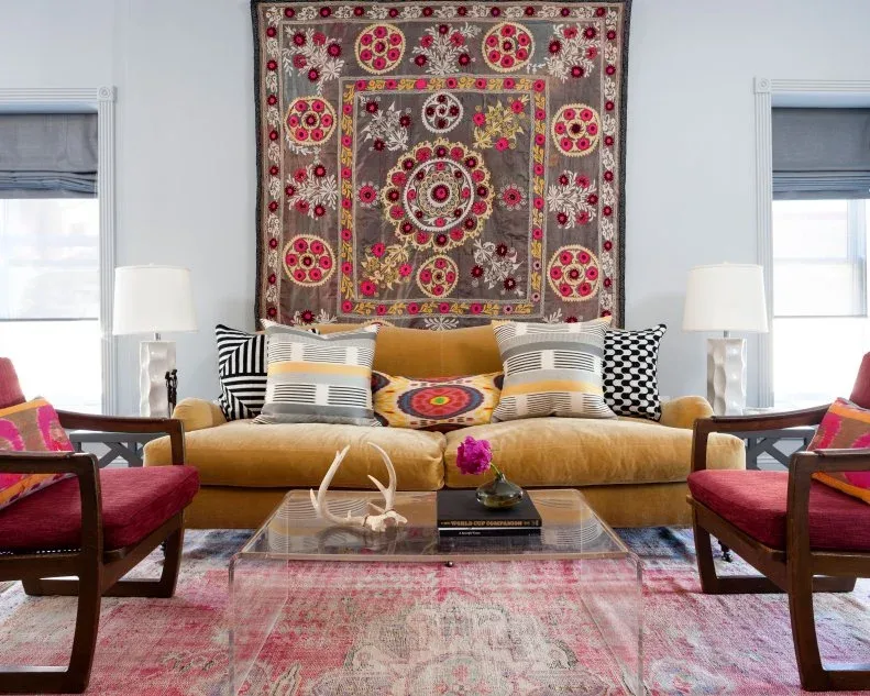 Gold-colored sofa below a patterned wall textile with pink chairs, a pink rug and glass table.