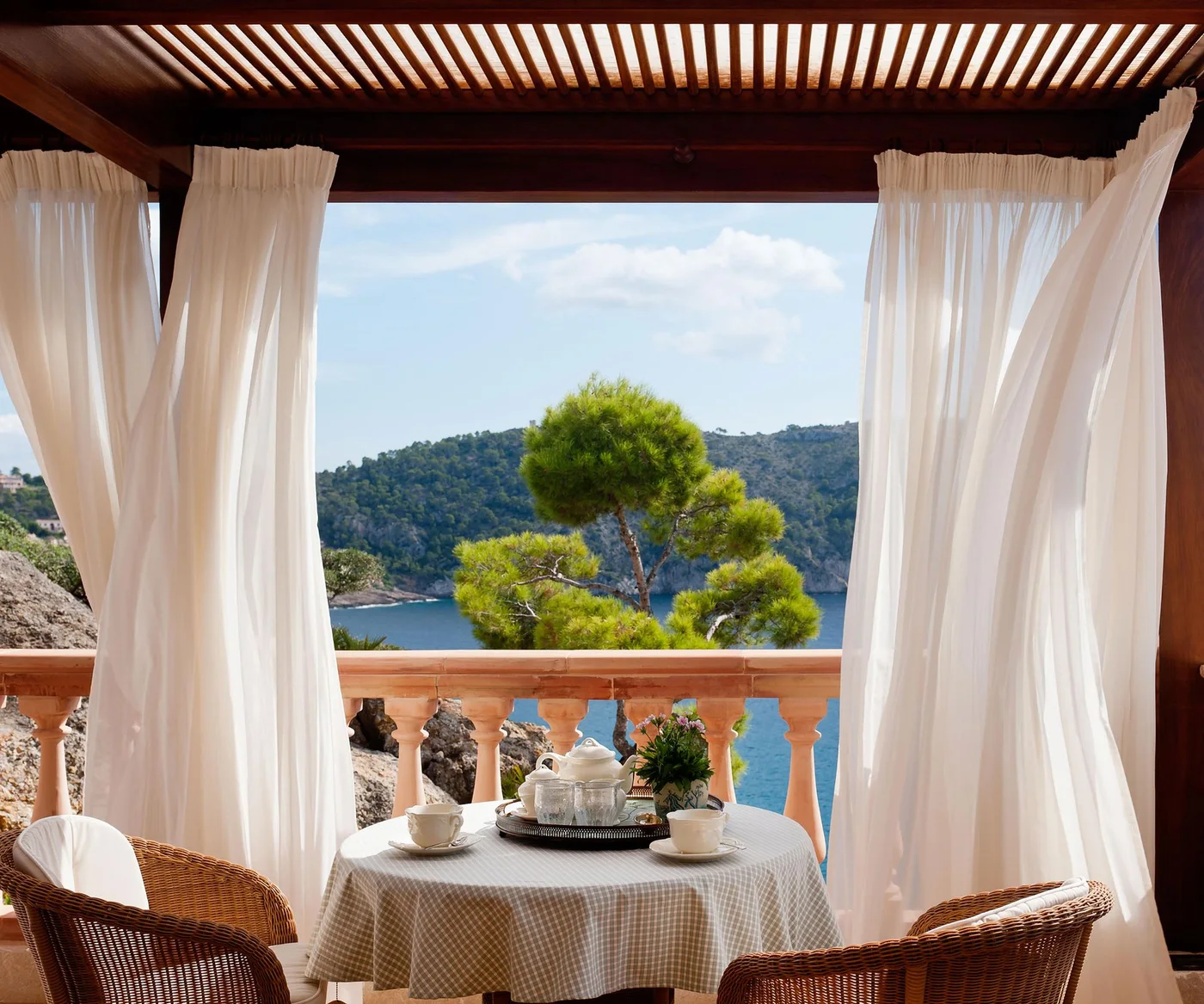 Pale curtains hang beneath a timber overhead frame around a small outdoor dining table