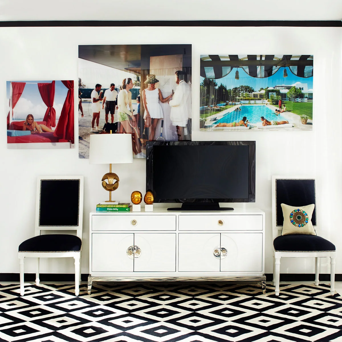 A television stands on a white console beside a gold-colored lamp, stacked books and small matching ornaments, with dark chairs at either side.