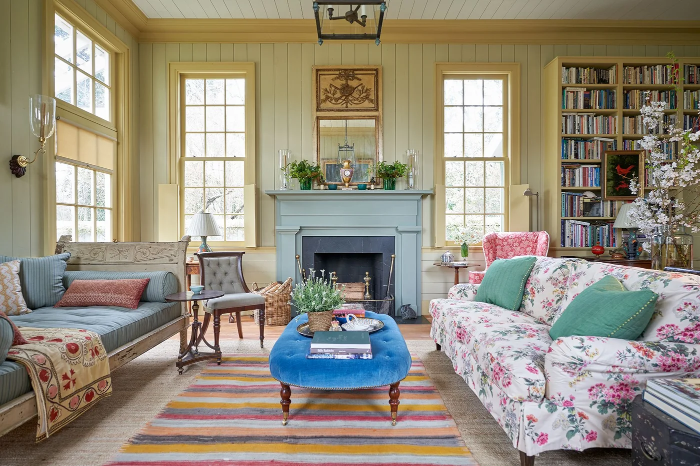 A muted blue mantel stands between tall windows in a pale yellow paneled room with floral seating and a blue ottoman.