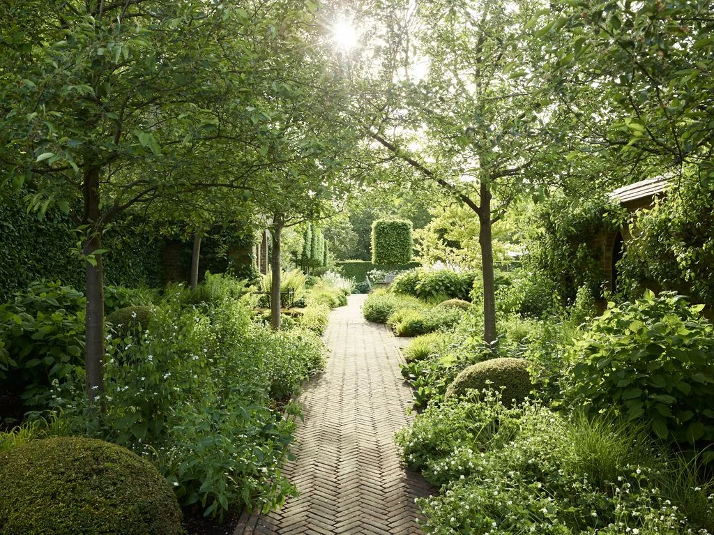 A herringbone brick path runs between planted borders toward a garden bench