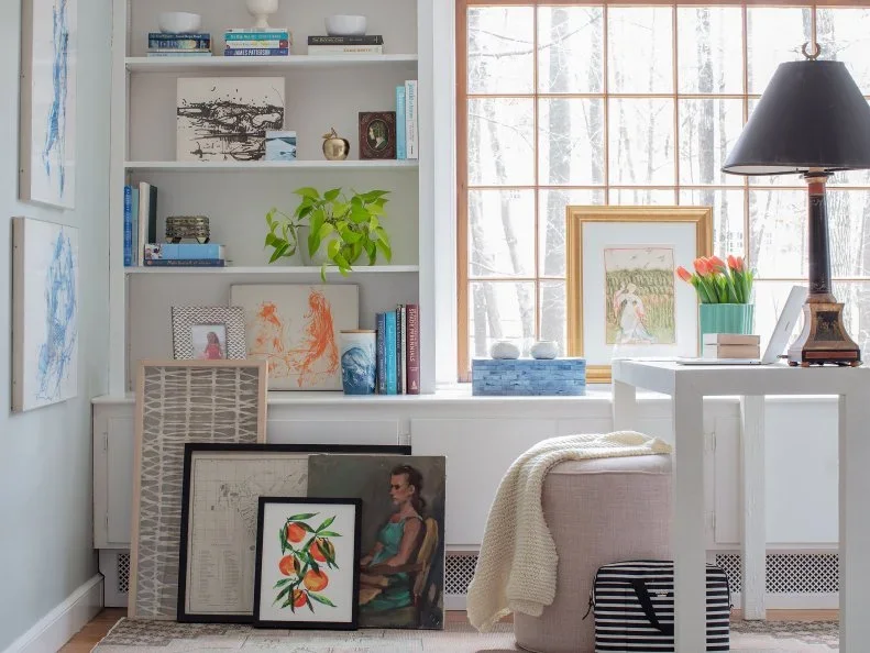 A slim pale desk stands beside built-in shelves and a large window, with a pale armchair, a dark-shaded lamp and books nearby.