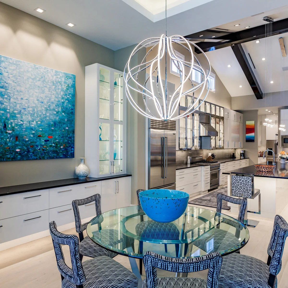 Large blue painting above white low cabinets beside a glass dining table in an open kitchen.