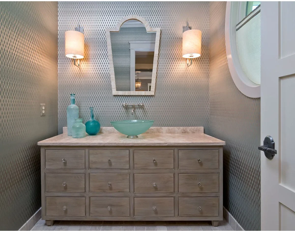 An aqua vessel bowl sits on a wide pale vanity with many drawer fronts beneath patterned gray walls and a shaped mirror.