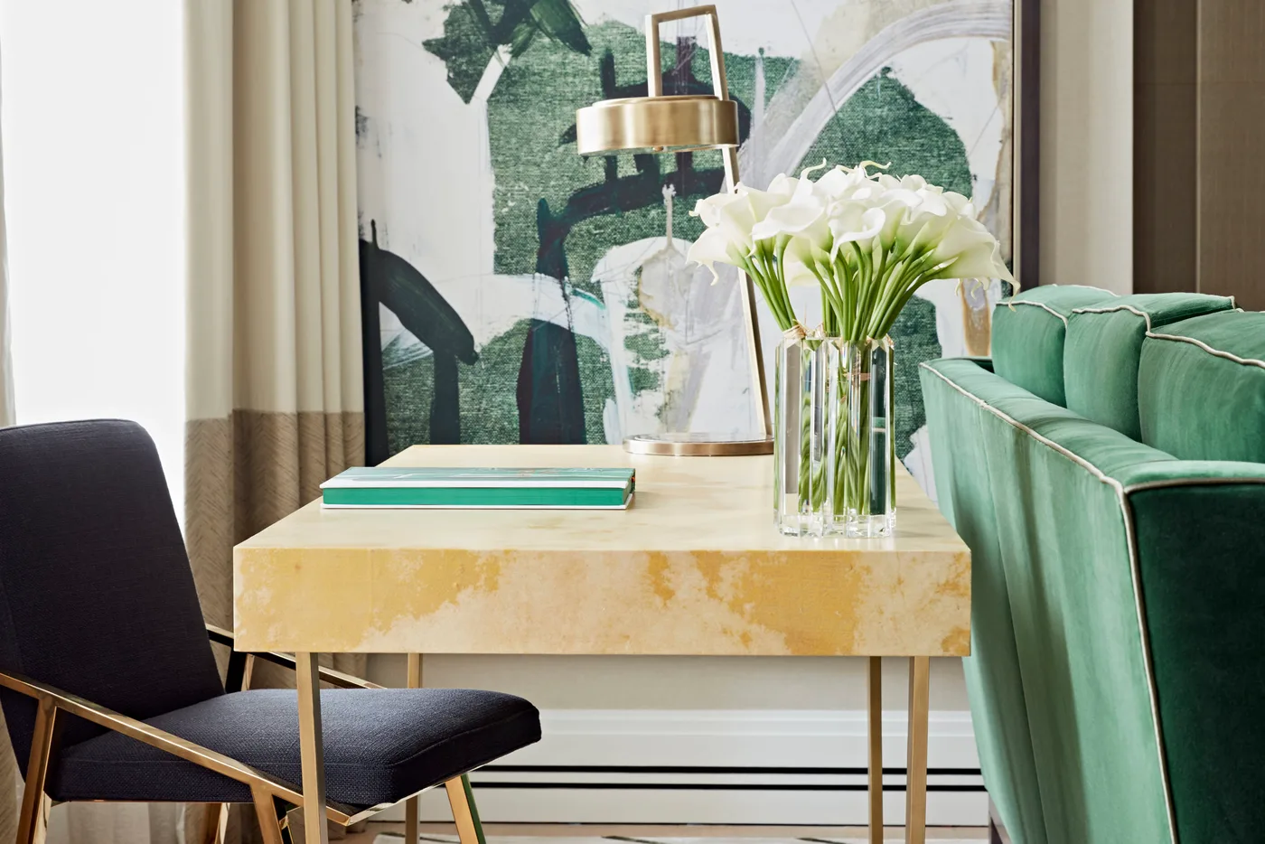 Writing desk with a gold-toned frame and lamp beside a green sofa and green abstract artwork.