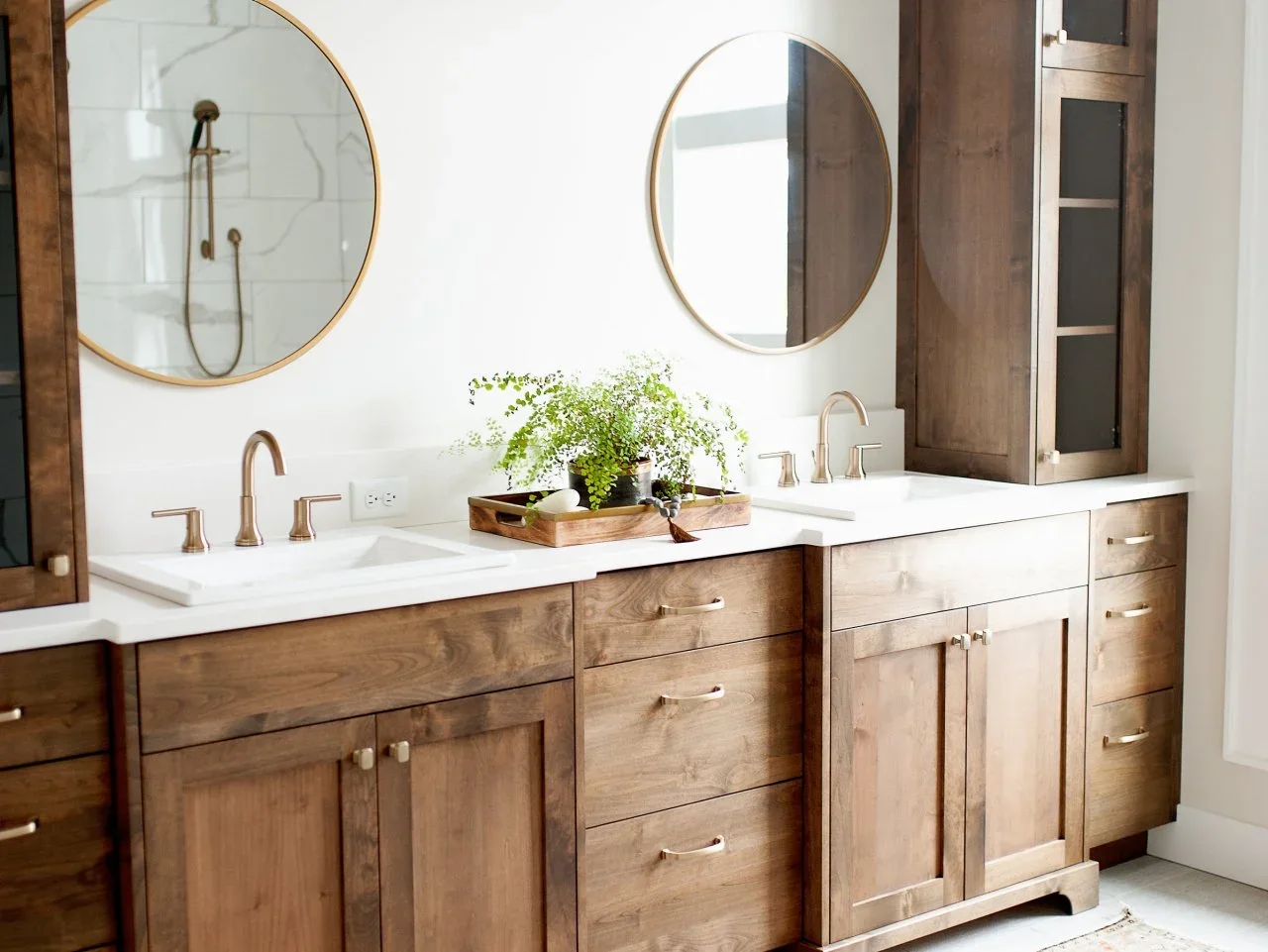 Timber vanity drawers and tall glass-front cupboards surround two white sinks and round mirrors.