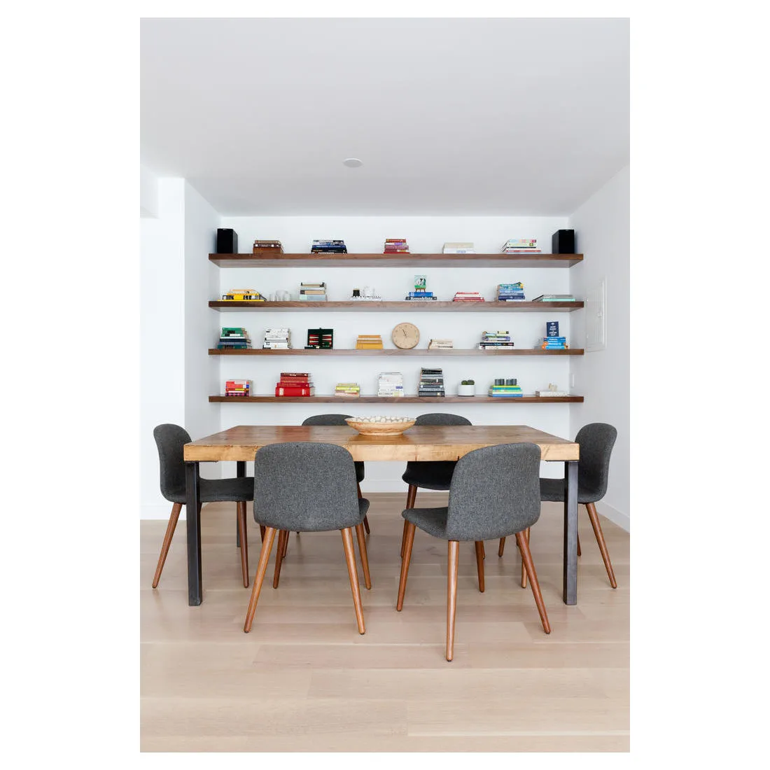 Four long wood shelves with books in a pale dining alcove behind gray chairs and a wood table