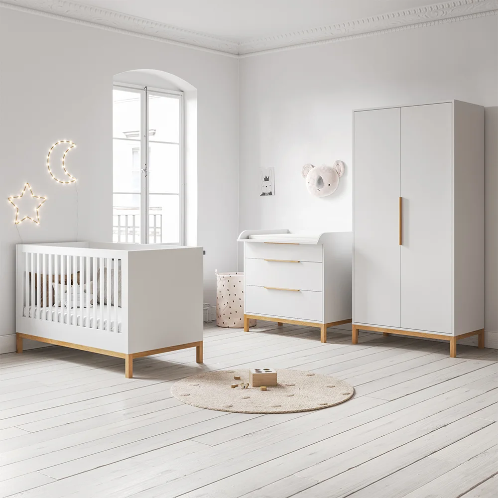 A white crib, drawer chest and tall wardrobe with wood legs stand beside pale walls, a broad window and a small round nursery rug
