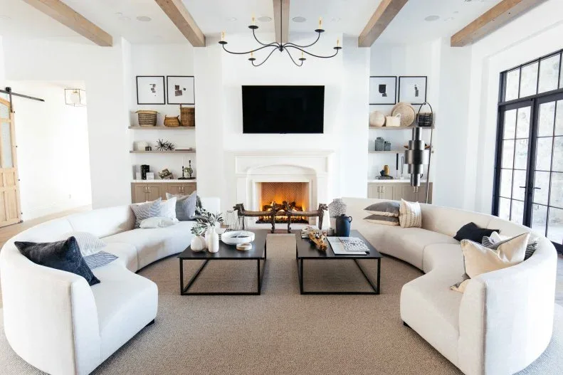 Living room with two pale curved sofas, separate black rectangular tables, white fireplace, open shelves, wooden ceiling beams and tall black-framed windows.