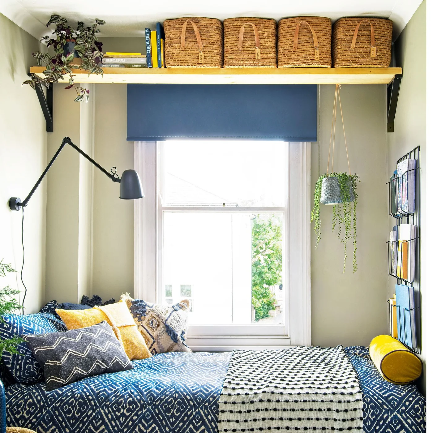 A narrow bed nook has blue patterned bedding, yellow cushions, a blue blind, a wall light and baskets on a high shelf above the window.