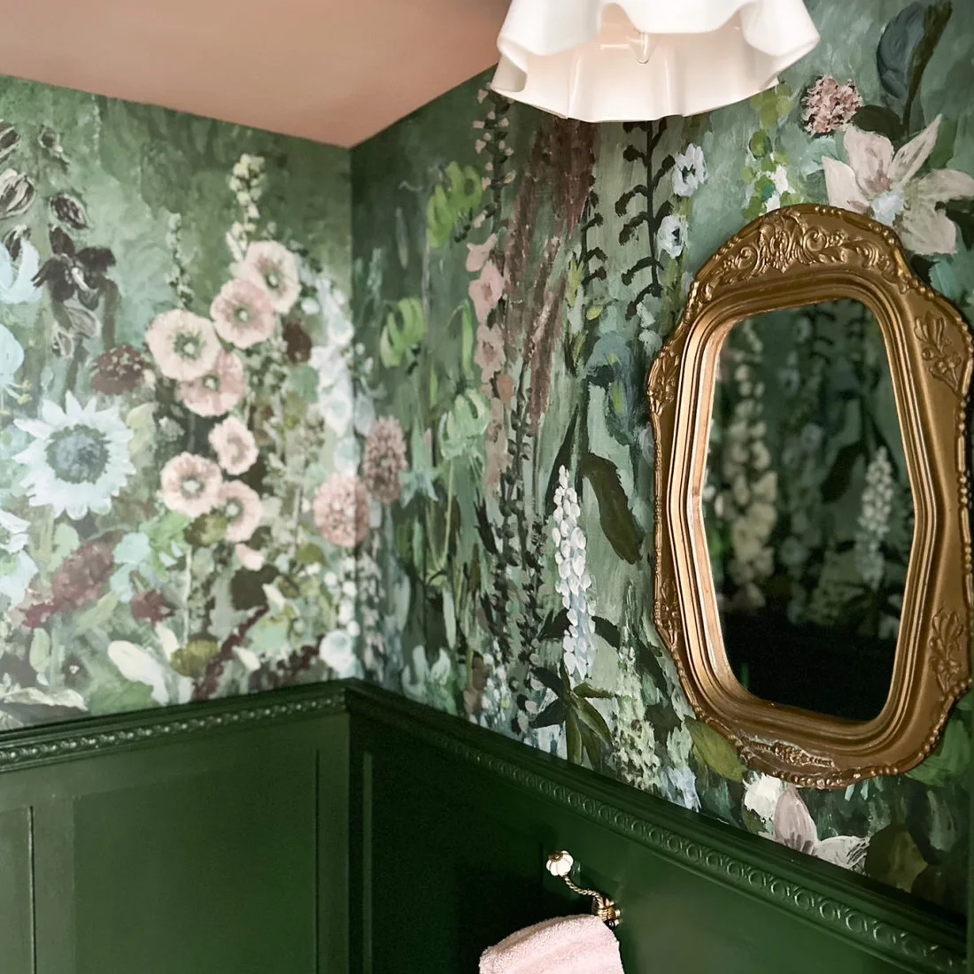 Large pale flowers cover green powder-room walls above dark green paneling beside an ornate gold-colored mirror and pale folded ceiling light