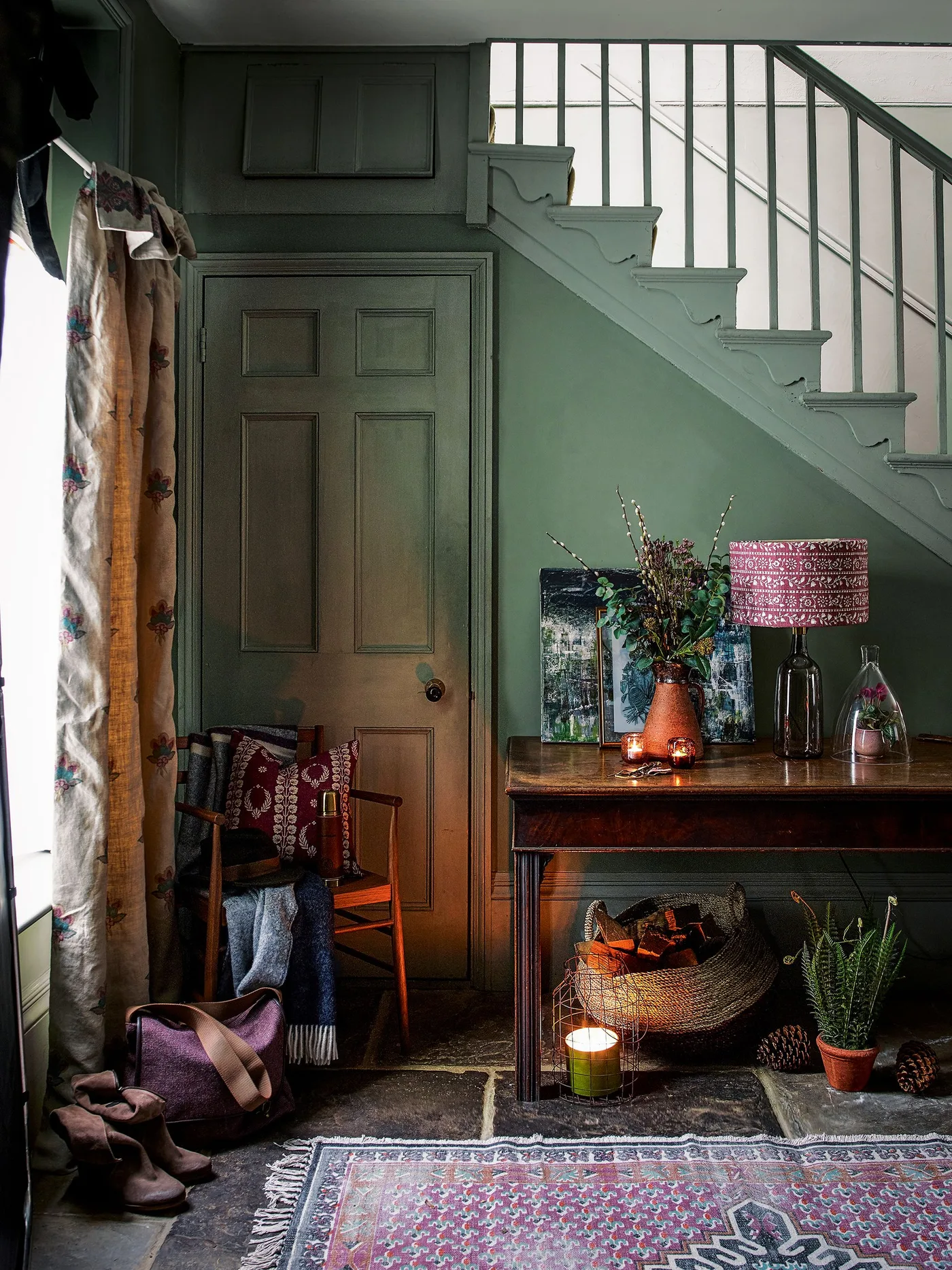 Muted pink and blue patterned rug in a green entry with wood console, chair, woven basket and pink-shaded lamp