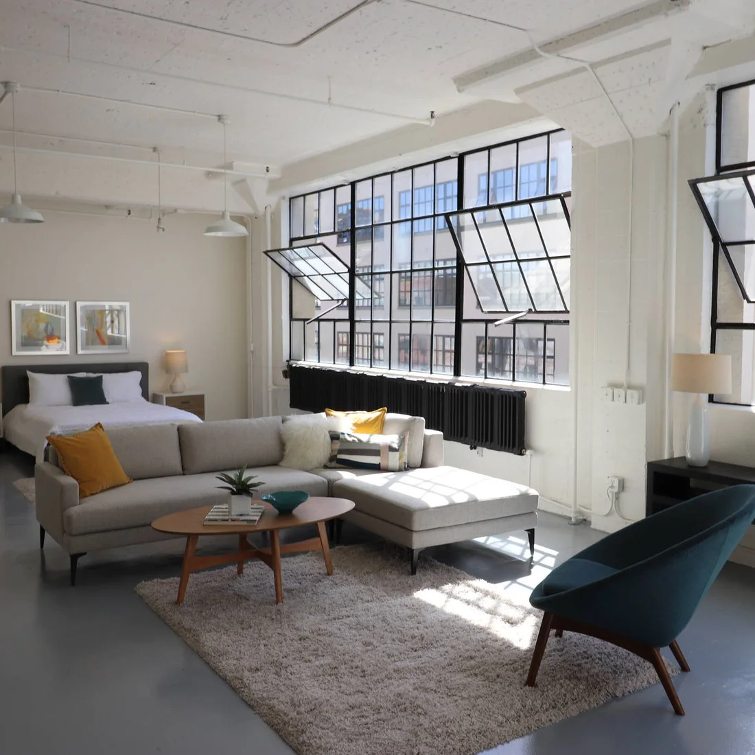 A gray chaise sofa and blue chair on a pale rug beside factory windows in an open studio