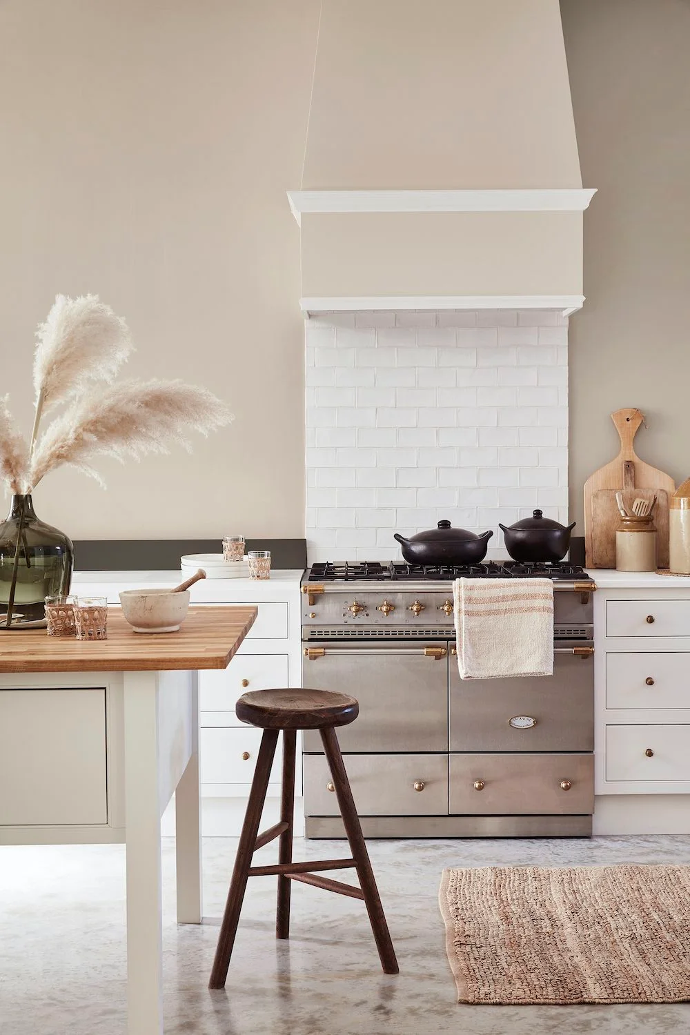 Soft-white drawers and an island sit beside a stainless range beneath a beige hood and pale rectangular tiles.