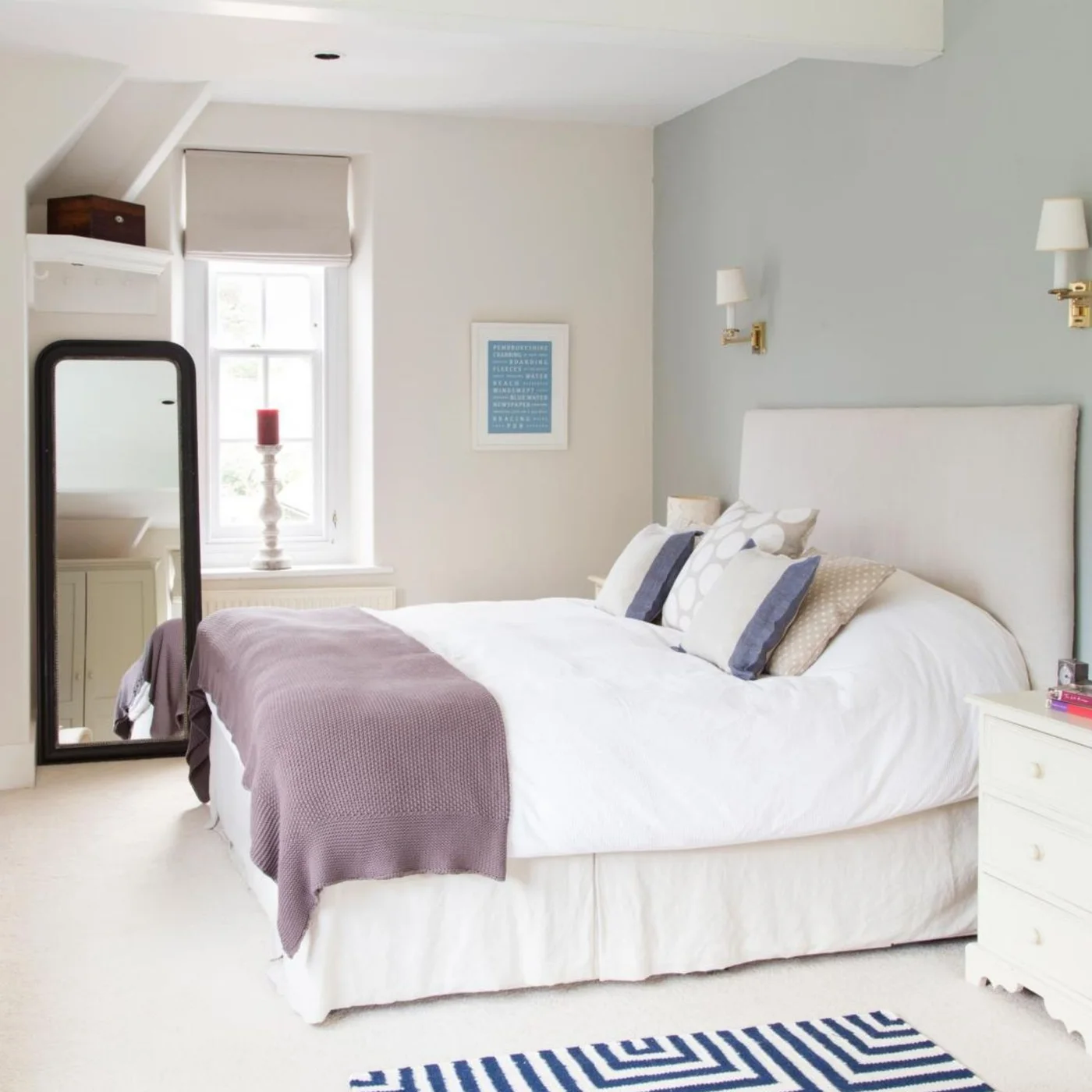 A pale bedroom has a tall dark-framed mirror in a recess beside the window, a white upholstered bed, small drawers and paired wall lights.