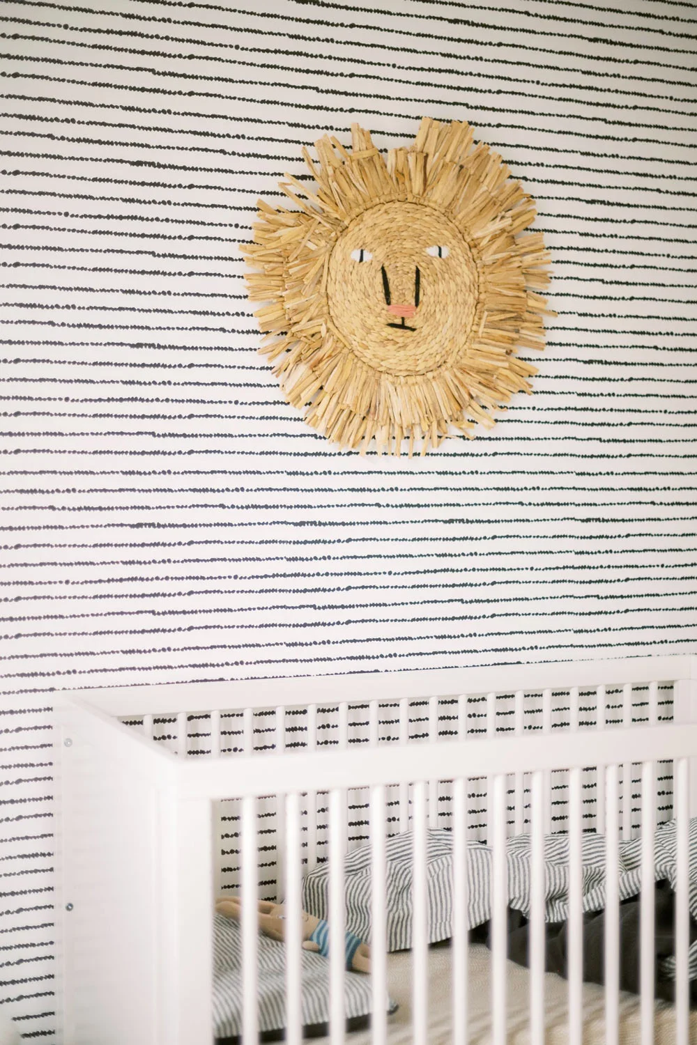 A warm sun-face wall ornament sits against fine dark horizontal nursery wallpaper above the edge of a white crib