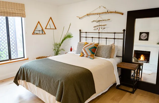 A dark metal bed with pale bedding and green throw stands beneath branch-like wall decoration beside small triangular mirrors, a woven shade and a dark-framed floor mirror