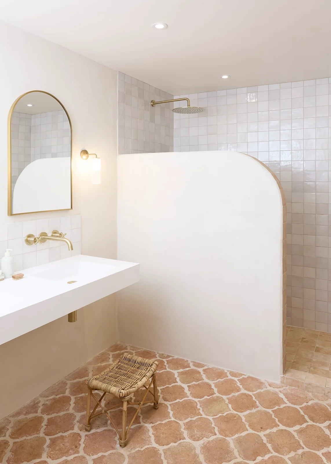 A compact pale bathroom has a curved built-in shower partition, rectangular wall-hung basin, arched mirror, warm-metal tap and peach-toned patterned floor.