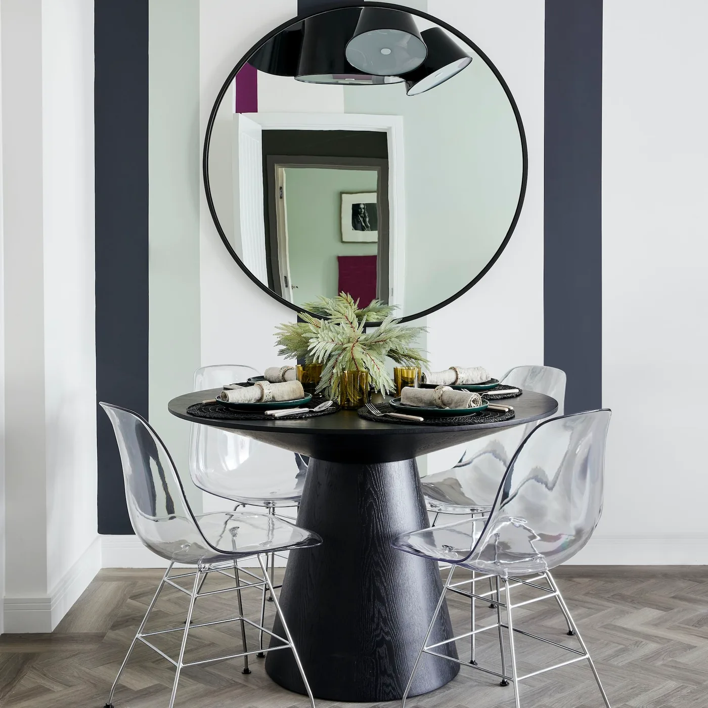 Large black round mirror above a small black round dining table with clear chairs and dark wall stripes