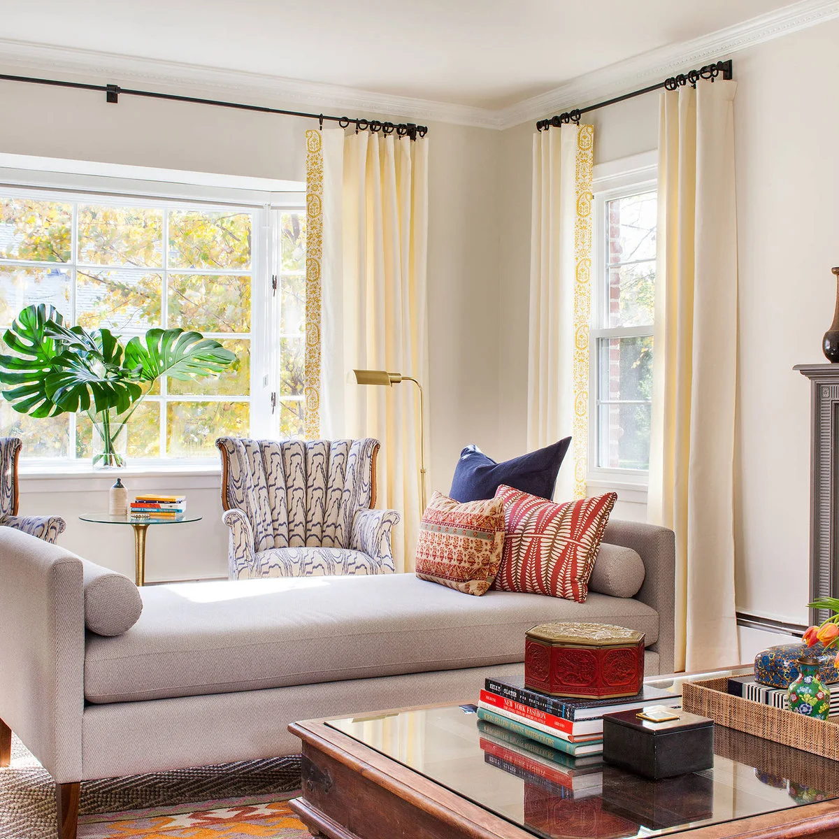 A pale gray daybed with raised ends sits beside patterned chairs, cream curtains and a low wood-and-glass table.