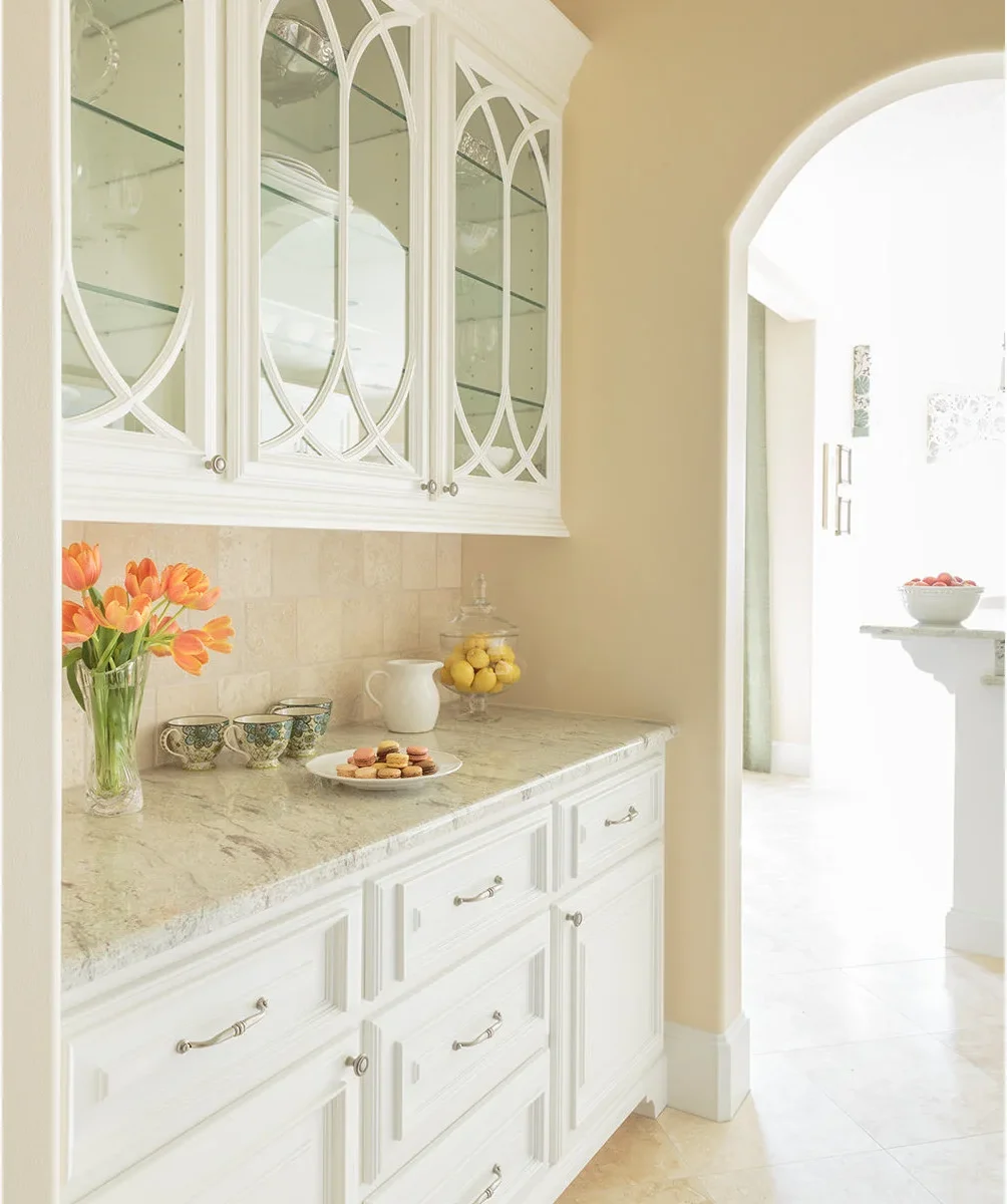 White glass-front cabinets with curved decorative frames sit against warm pale yellow walls above a pale patterned counter.