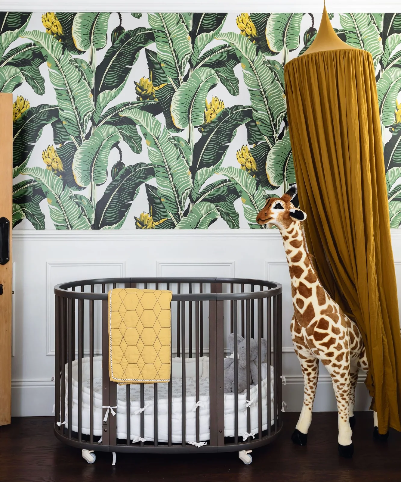 Oversized green banana-leaf wallpaper rises above white paneling beside a dark oval crib, yellow patterned blanket and yellow canopy