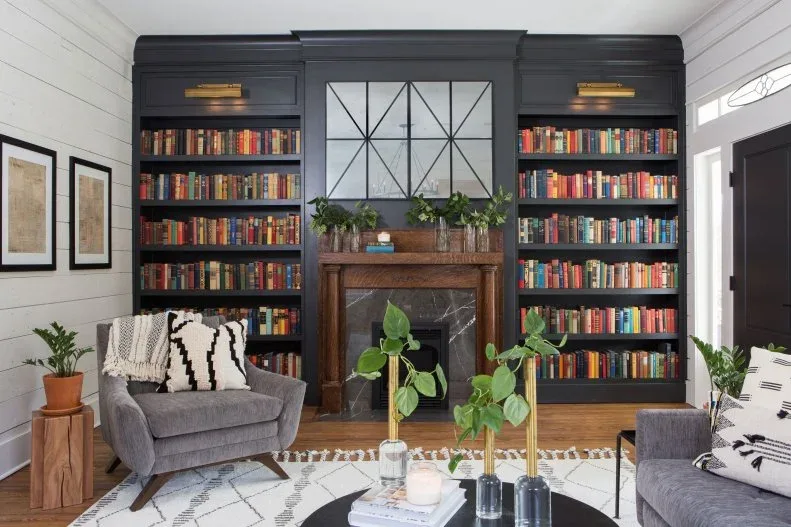 Tall dark bookcases with brass-toned lights around wood fireplace mantel and divided mirror