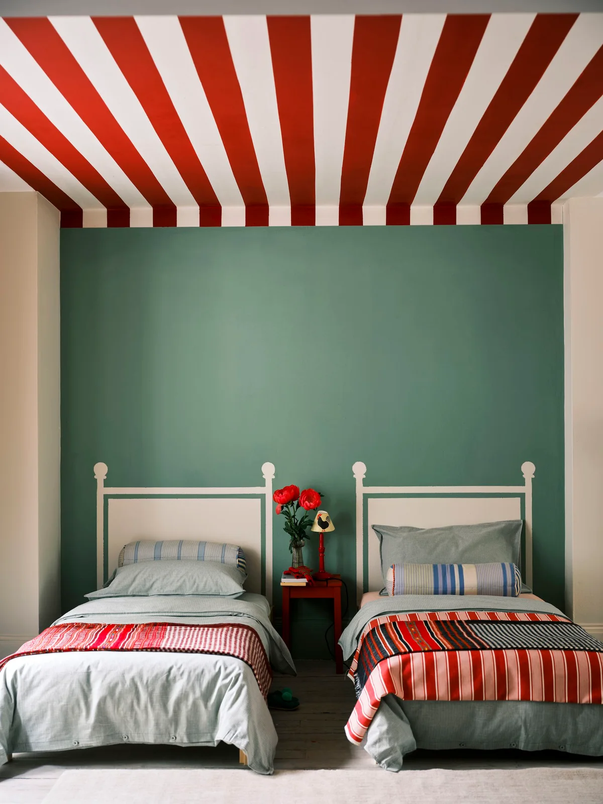 Two pale metal-frame beds sit beneath a red-and-white striped ceiling beside green walls, patterned pillows and a small red central table