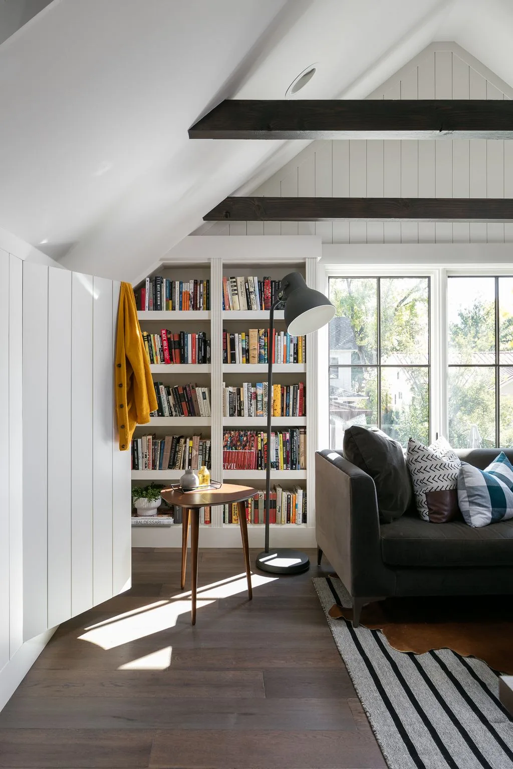 Living room corner with white paneling, dark ceiling beams, a tall bookcase, dark sofa and a reading lamp.