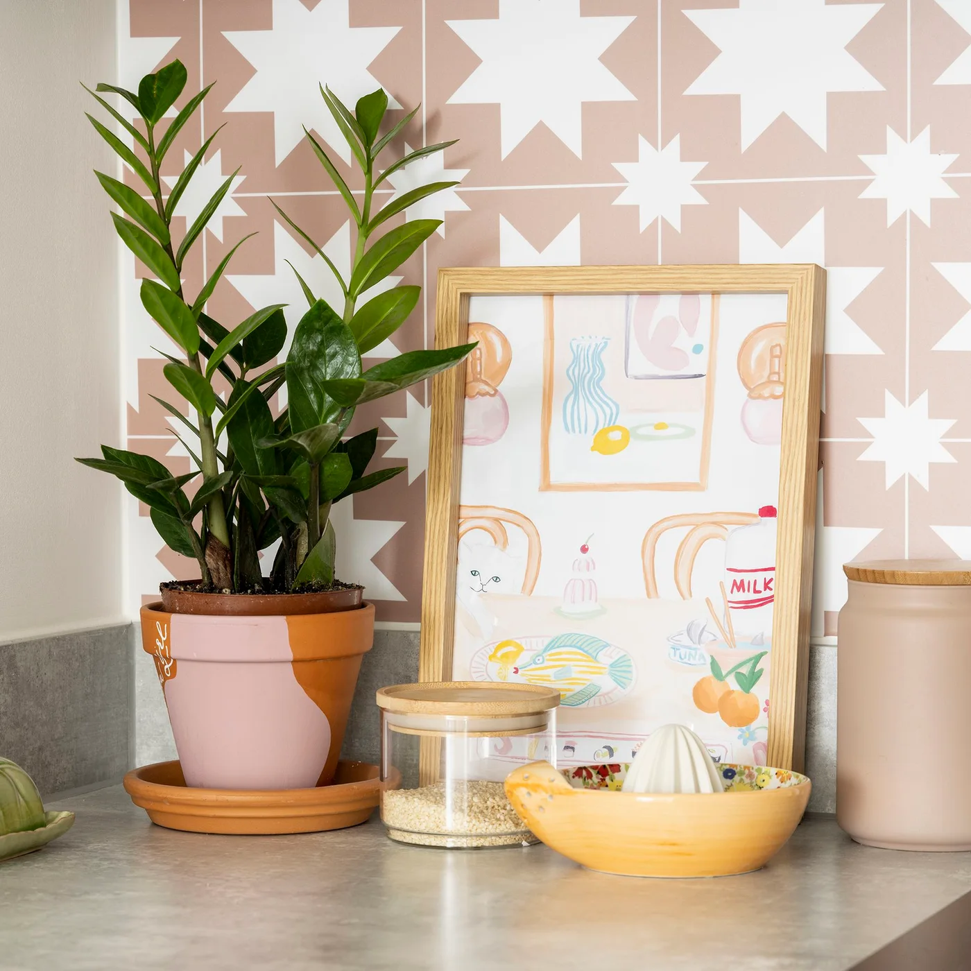Pink-and-white geometric tiles sit behind a kitchen counter with a plant, framed print, canisters and a small citrus press