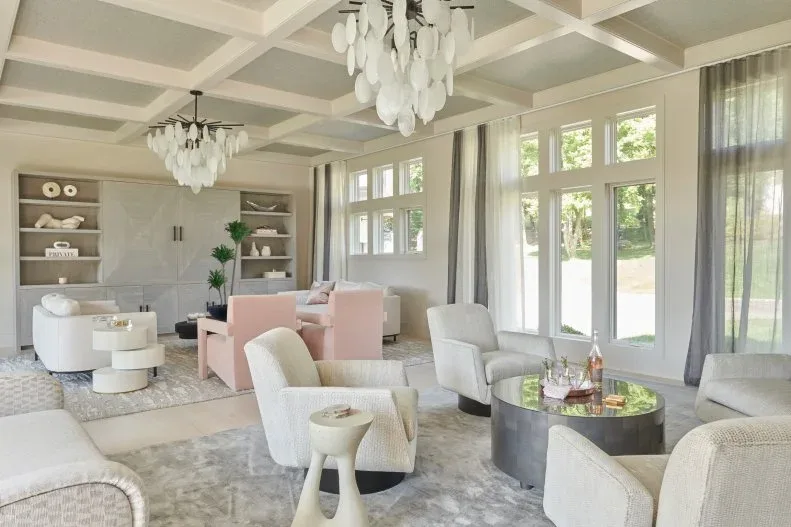 Large living room with rounded pale chairs, pale pink chairs farther back, circular tables, gray rugs, coffered ceiling and decorative white pendant lights.