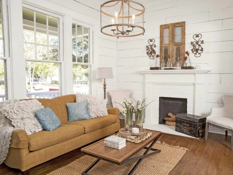 A white shiplap living room has a pale mantel and white brick fireplace, mustard sofa, small wood table and tall windows along the adjoining wall.