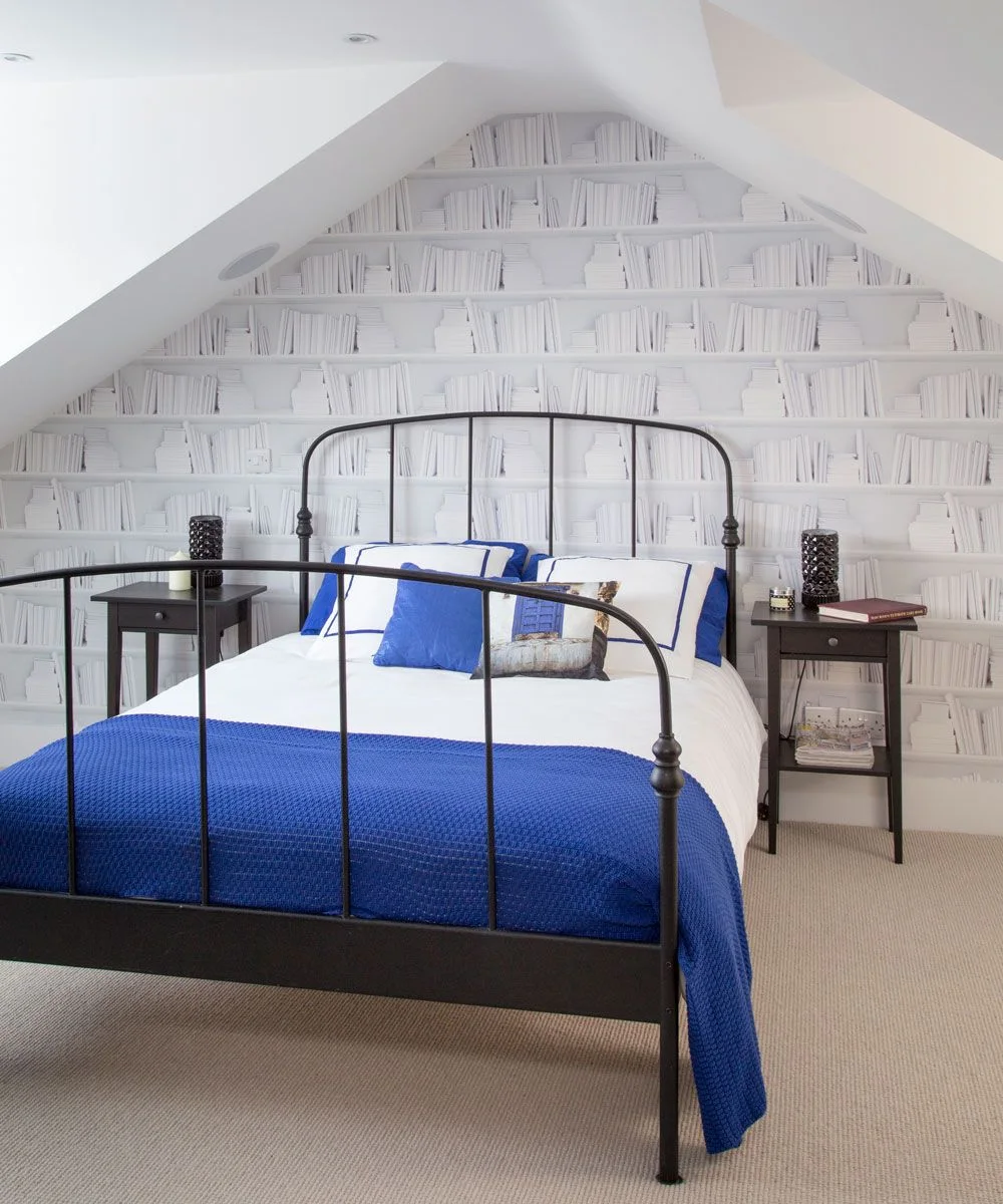 Bookcase-pattern wallpaper on an attic gable behind a black metal bed with blue-and-white bedding.