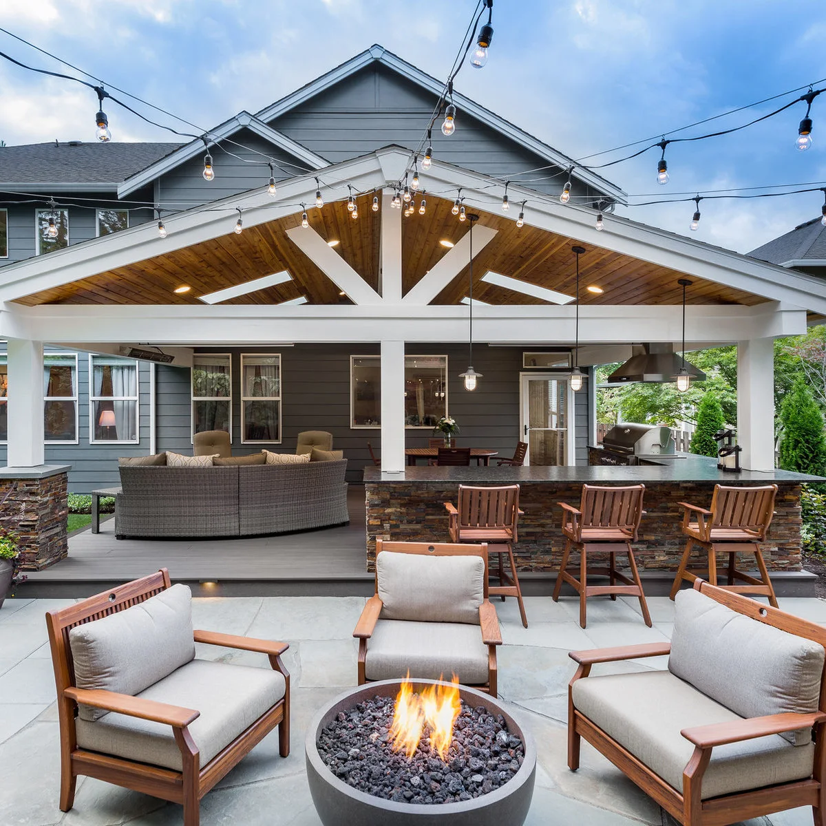 A broad white gabled roof shelters lounge seating, dining and a counter beside an open fire-pit terrace.