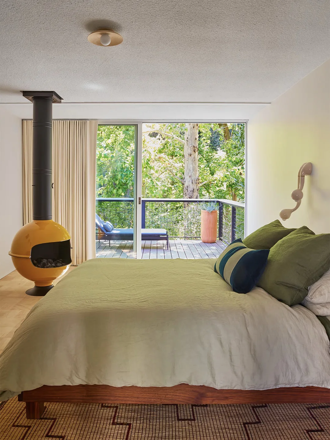 A sculptural yellow bedroom fireplace stands beside pale green bedding and a wood bed frame near tall curtains and a garden-facing glazed door