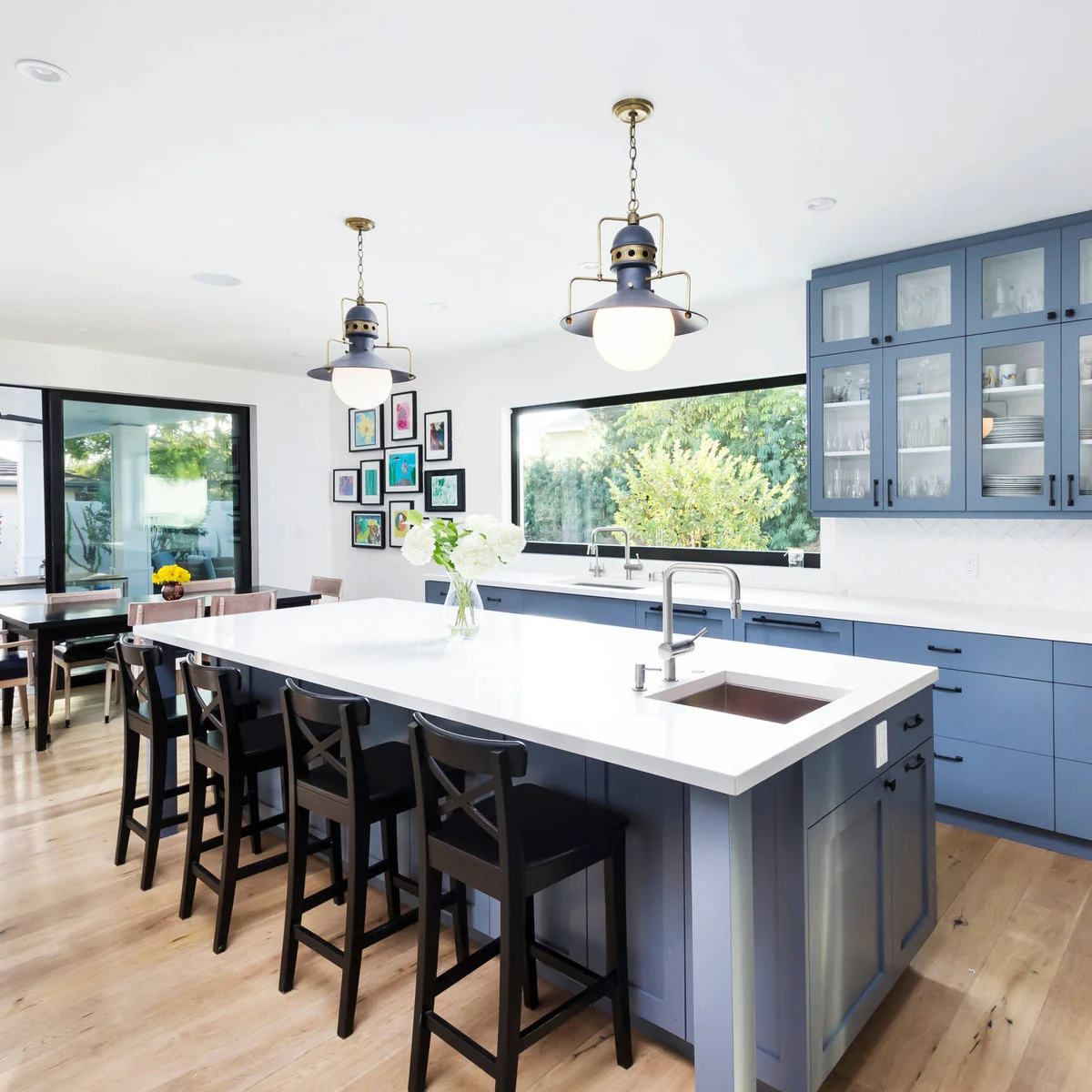 A long dark-framed kitchen window above a sink with blue cabinets and a small gallery of colorful art to the side.