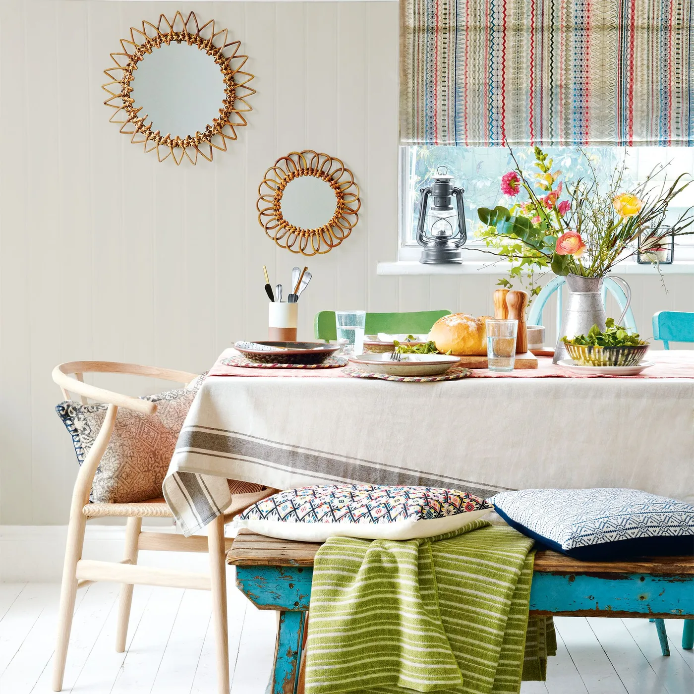 Two round woven-frame mirrors beside a striped blind above a colorful dining table and turquoise bench
