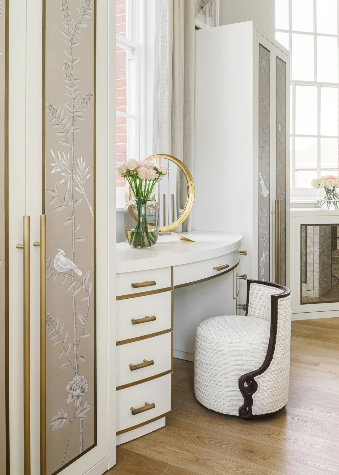 A curved pale dressing surface with drawers fits between decorated wardrobe fronts beside a rounded upholstered seat