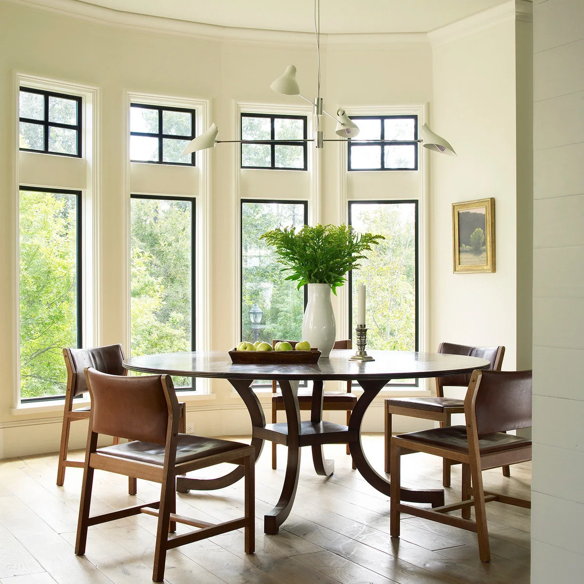 A round dark table with wood-framed brown chairs in front of a broad curved row of tall windows.