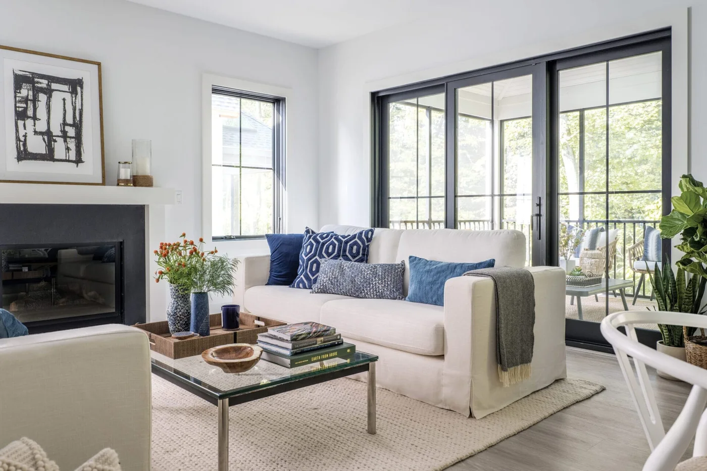 A white sofa with blue cushions faces a glass coffee table beside dark-framed windows, a dark fireplace opening and a light woven rug