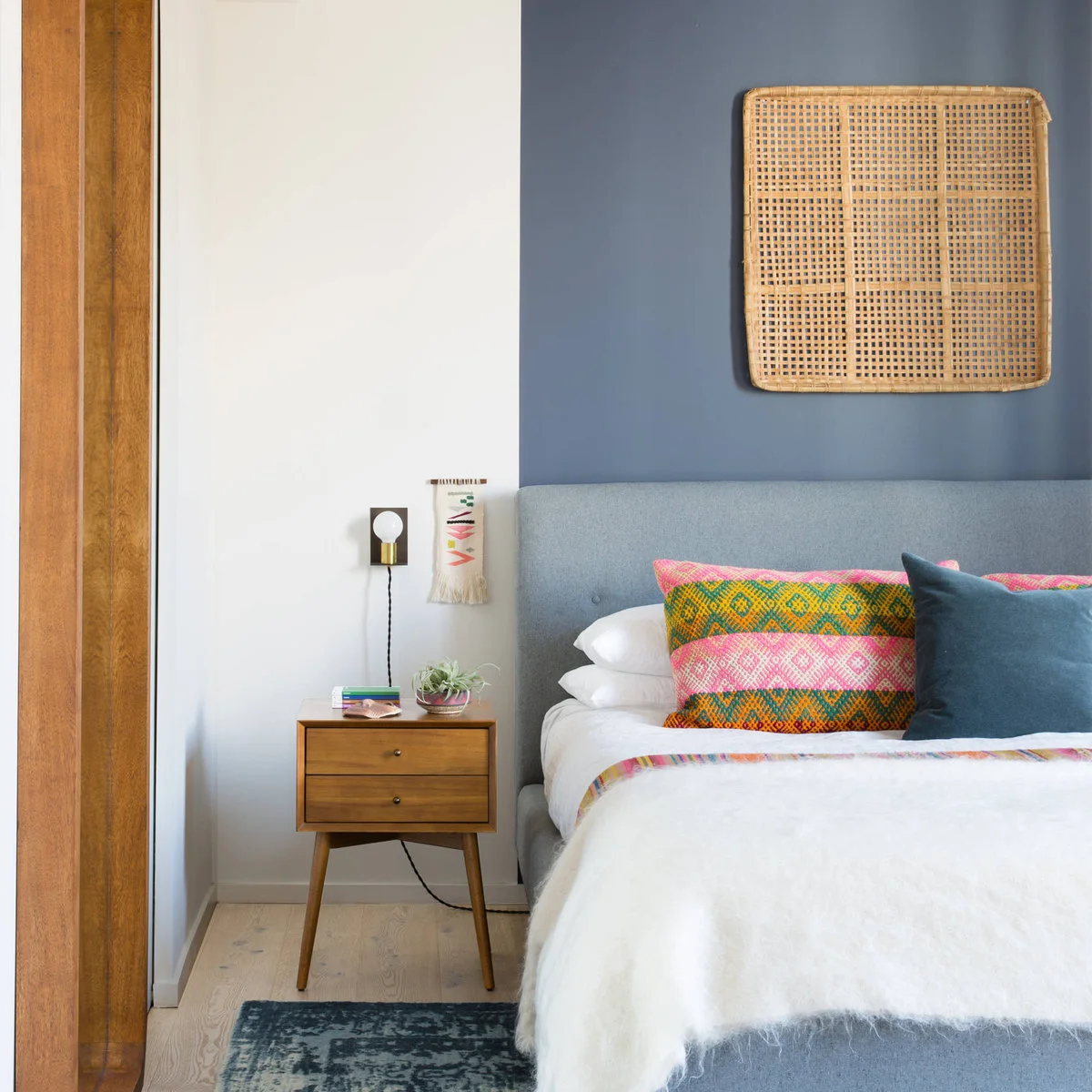 Small wood-toned two-drawer nightstand beside a blue upholstered bed and wall bulb