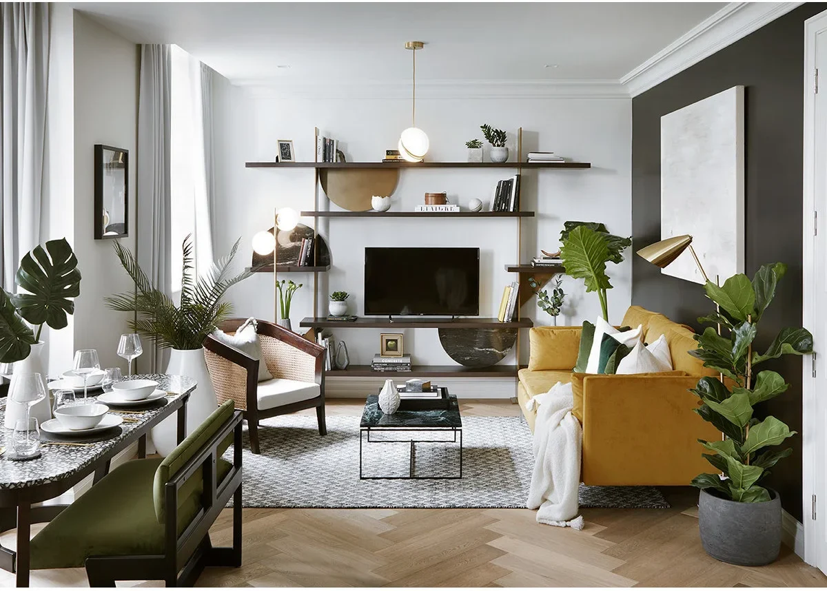 A mustard sofa and compact dark table face a television on open wall shelves, with a dining table and green chairs along the window side.
