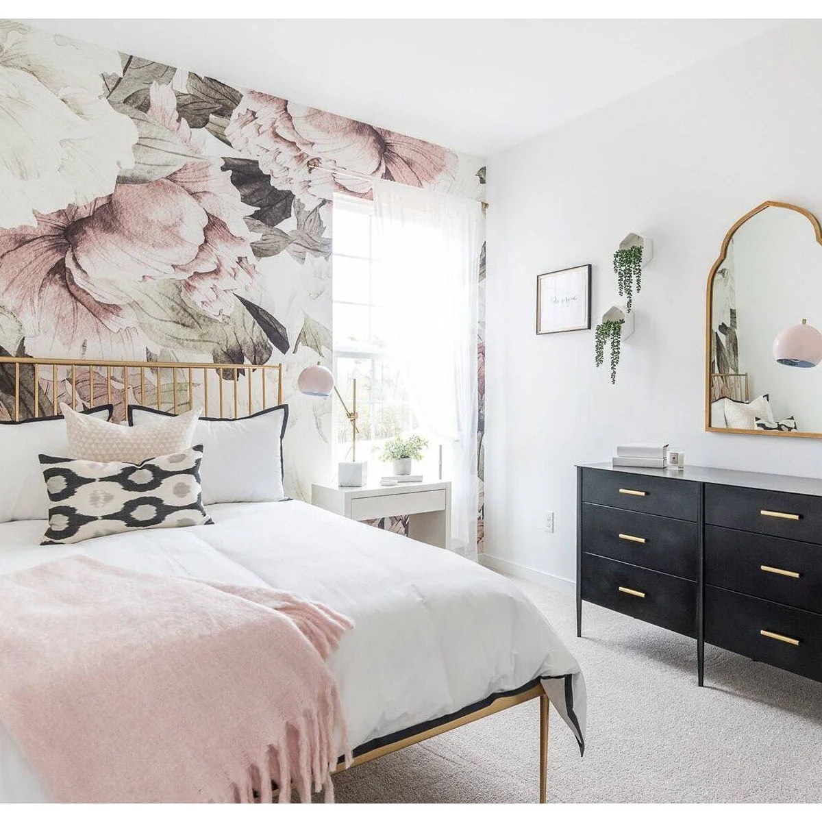 Pale pink floral mural behind a slim gold-colored bed with white bedding, pink lamps and a black dresser.