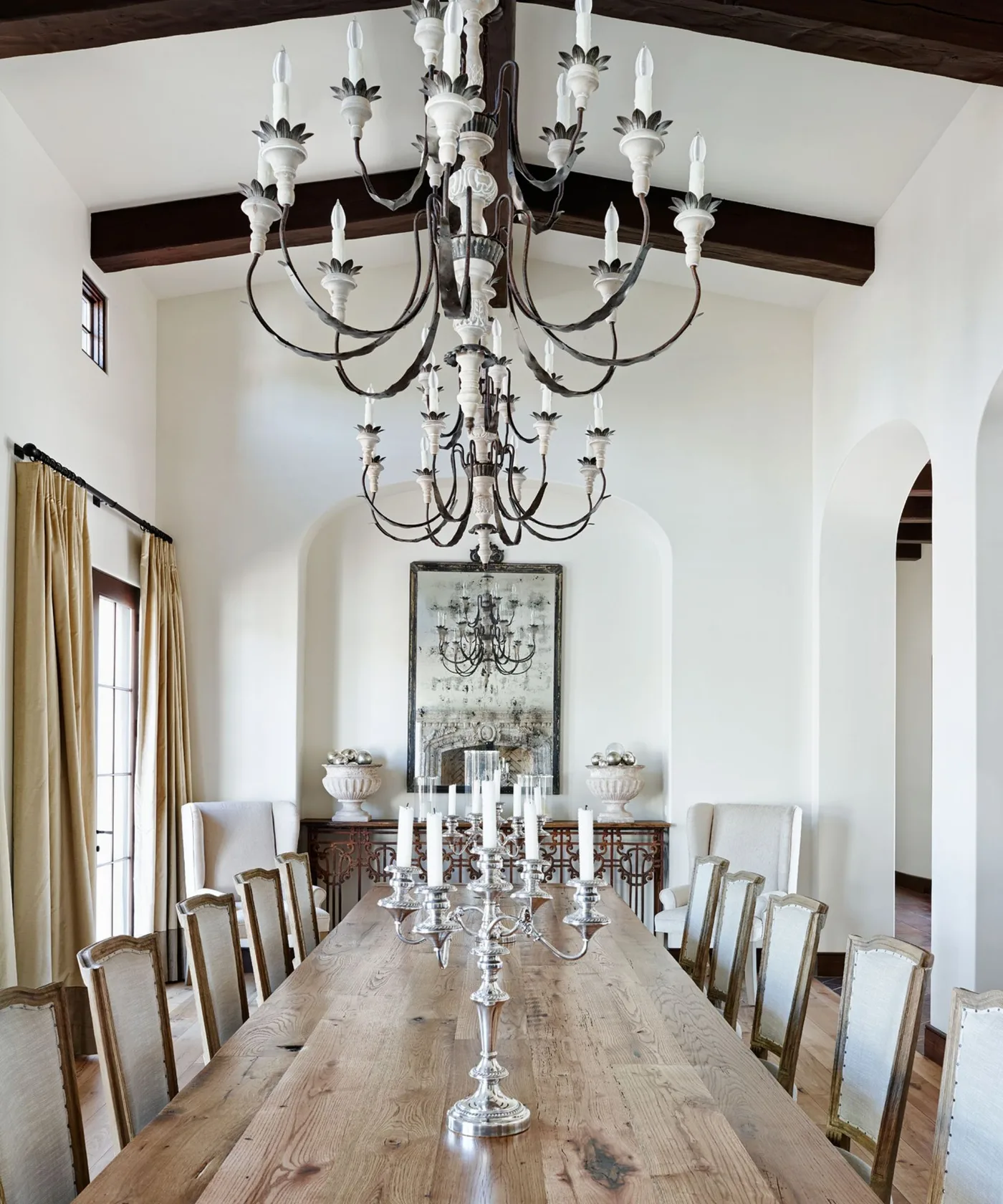 Long rustic dining table with light wooden chair backs, exposed dark beams and two decorative chandeliers.
