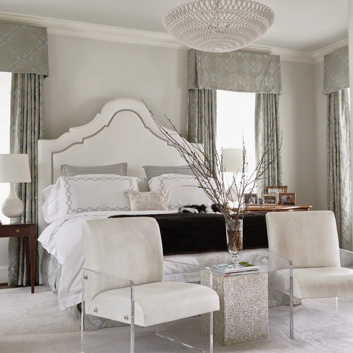 Pale bedroom with a high curved white headboard, patterned curtains and two cream chairs