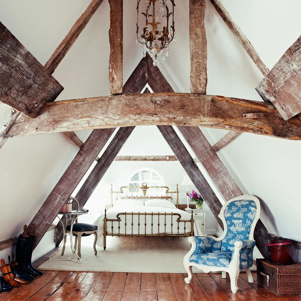 Long attic bedroom with exposed wooden beams, a metal bed, a blue patterned chair and pale walls.